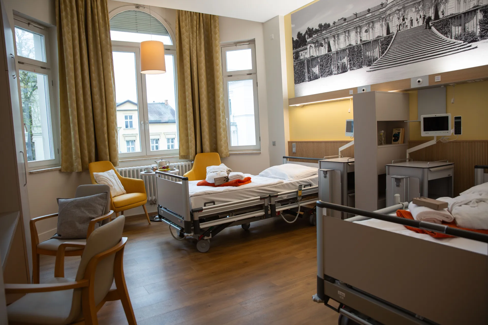 Two hospital beds in a bright patient room with large windows, yellow curtains and a wooden floor
